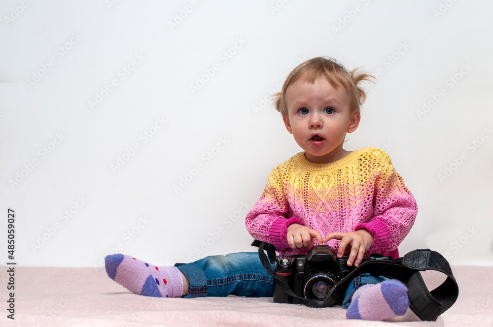 The child plays with the camera. A little one-year-old girl with blond hair sits on a bed and looks at a SLR camera. The child plays with the camera. A little one-year-old girl with blond hair sits on a bed and looks at a SLR camera.