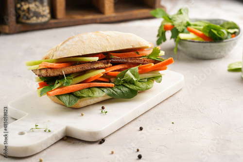 Traditional vietnamese banh mi sandwich with sliced smoked tofu, fresh spinach, shredded carrots and peeled cucumbers, cilantro on marble board on light grey textured background