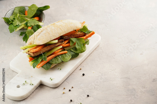 Traditional vietnamese banh mi sandwich with sliced smoked tofu, fresh spinach, shredded carrots and peeled cucumbers, cilantro on marble board on light grey textured background, copy space photo
