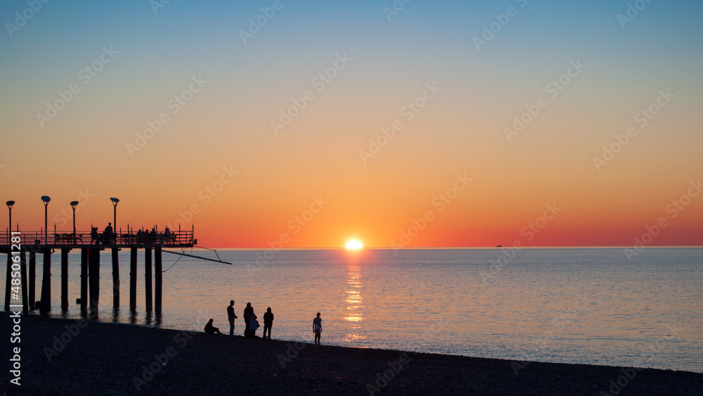 Colorful seascape, sunset shot and red sky. Beach and pier with people silhouette in backlight, coastal sunlight on water