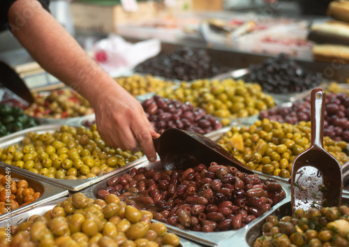Different colors and taste olives in plastic boxes on stall in foreground. Market. Seller's hand picks up brown olives with scoop. Traditional arabic and jewish food. Mediterranean appetizer. 