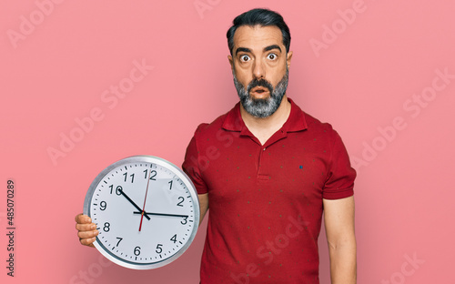 Middle aged man with beard holding big clock scared and amazed with open mouth for surprise, disbelief face