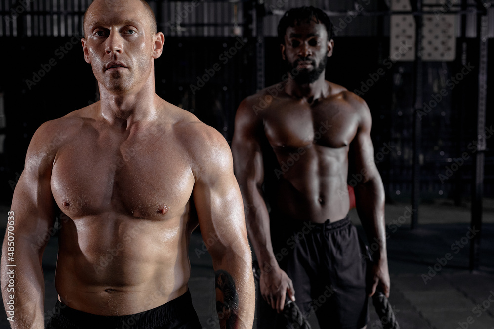 two Athletic young men with battle rope doing exercise in functional training fitness gym. shirtless muscular males holding battle ropes in hands, exercising. side view portrait. cross fit concept two Athletic young men with battle rope doing exercise in functional training fitness gym. shirtless muscular males holding battle ropes in hands, exercising. side view portrait. cross fit concept