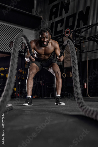fit active afro man with battle rope doing exercise in functional training fitness gym exercising with battle ropes at gym. black shirtless guy working out arms and cardio for cross fit