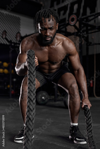Young black man doing strength training using battle ropes at modern dark gym. Athlete moving the ropes in wave motion as part of fat burning workout, active intense cross fit training, exercising