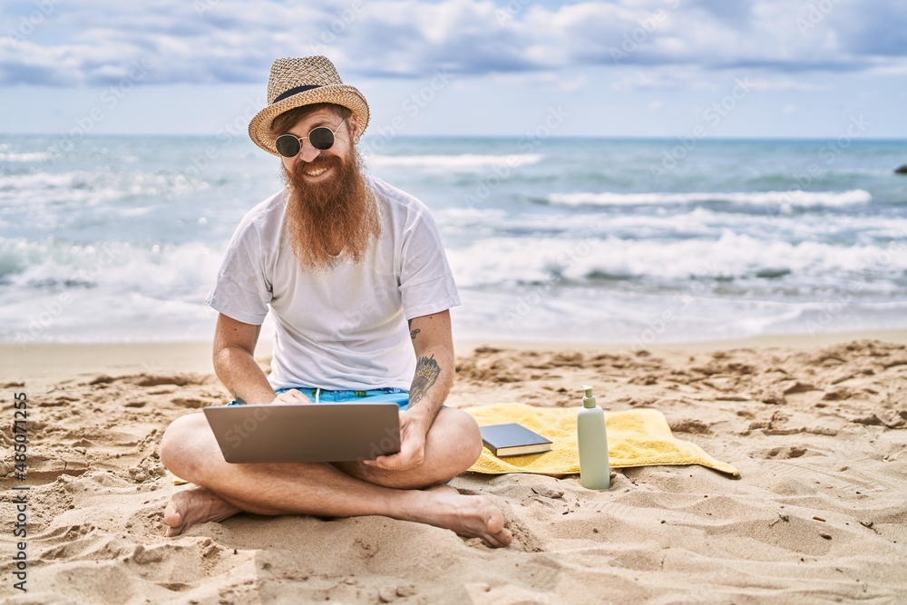 Young redhead man using laptop sitting on the towel at the beach.