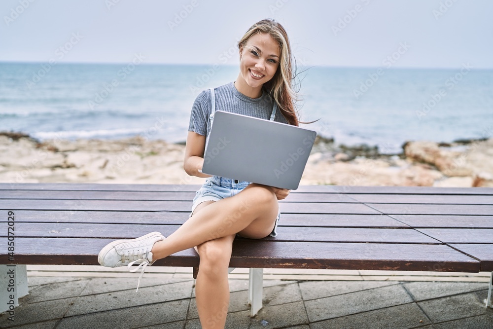 Young caucasian girl smiling happy using laptop sitting on the bench at the beach.