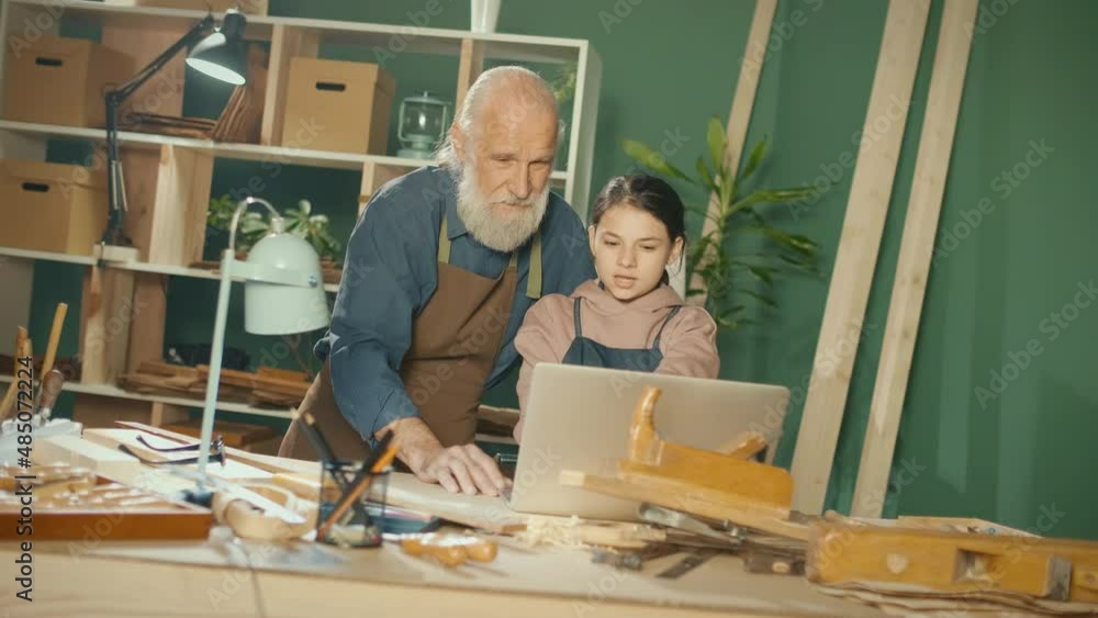 Grandfather Carpenter with his Teenage Granddaughter Develop Wood Layouts, Model 3D Sketches Using a Laptop in the Workshop. Communication of Generations. Craft, Joinery and New Computer Technologies. Grandfather Carpenter with his Teenage Granddaughter Develop Wood Layouts, Model 3D Sketches Using a Laptop in the Workshop. Communication of Generations. Craft, Joinery and New Computer Technologies.
