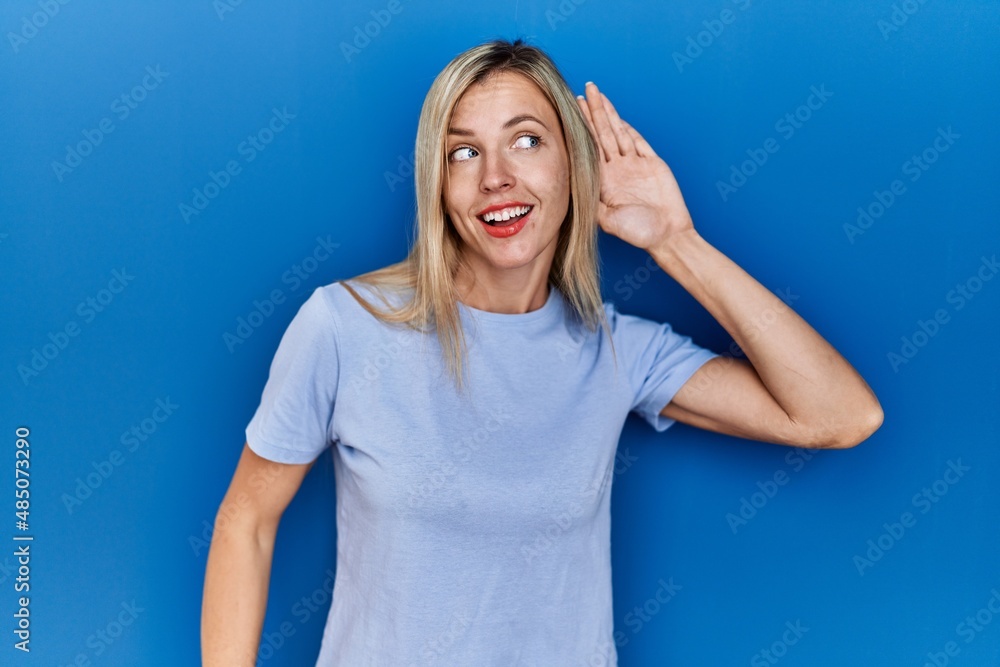 Beautiful blonde woman wearing casual t shirt over blue background smiling with hand over ear listening an hearing to rumor or gossip. deafness concept. Beautiful blonde woman wearing casual t shirt over blue background smiling with hand over ear listening an hearing to rumor or gossip. deafness concept.