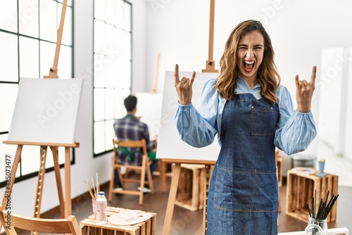 Young artist woman at art studio shouting with crazy expression doing rock symbol with hands up. music star. heavy concept.