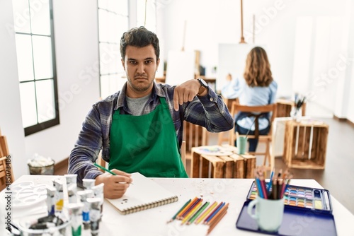 Young artist man at art studio pointing down looking sad and upset, indicating direction with fingers, unhappy and depressed.