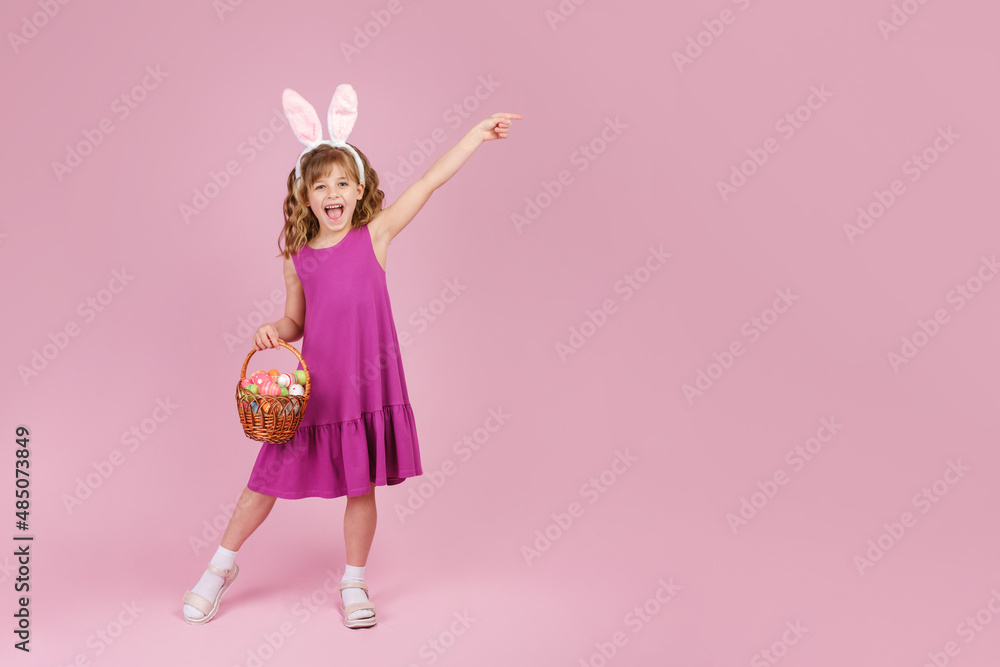 Full body little beautiful smiling girl in Easter bunny ears holds a basket with eggs on pink background and pointing away with hand. Copy space for text