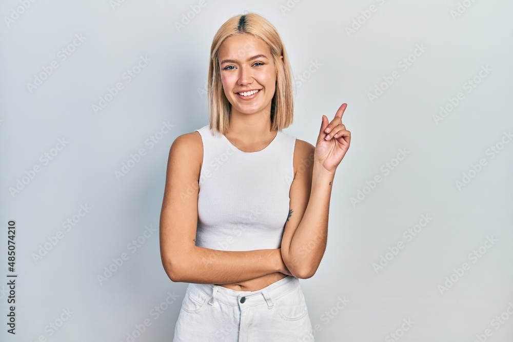 Beautiful blonde woman wearing casual style with sleeveless shirt with a big smile on face, pointing with hand and finger to the side looking at the camera. Beautiful blonde woman wearing casual style with sleeveless shirt with a big smile on face, pointing with hand and finger to the side looking at the camera.