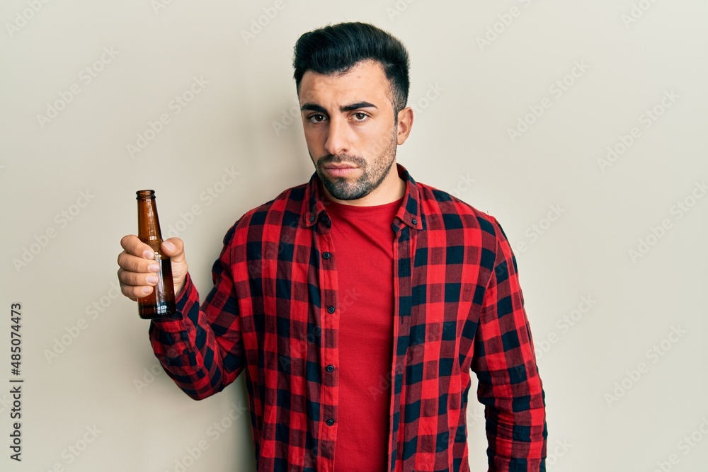 Young hispanic man drinking a bottle of beer thinking attitude and sober expression looking self confident Young hispanic man drinking a bottle of beer thinking attitude and sober expression looking self confident