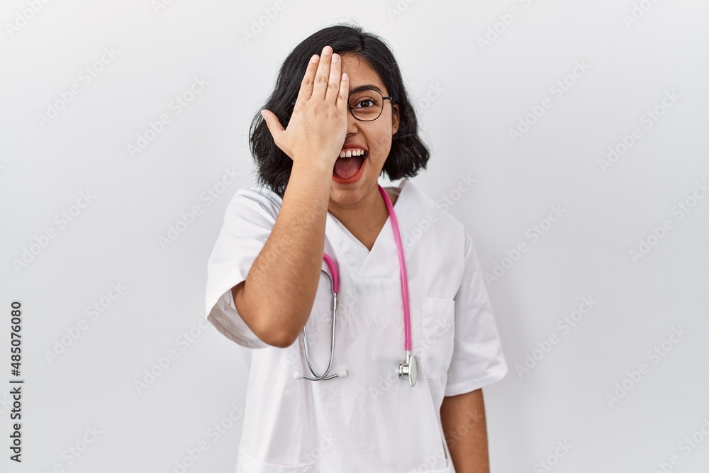 Young hispanic doctor woman wearing stethoscope over isolated background covering one eye with hand, confident smile on face and surprise emotion. Young hispanic doctor woman wearing stethoscope over isolated background covering one eye with hand, confident smile on face and surprise emotion.