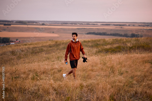 sportive man in hoodie walking in wheat field having rest after sport fitness, caucasian guy in casual hoodie alone at sunrise in the morning in mountains, nature landscape. jogging, sport concept