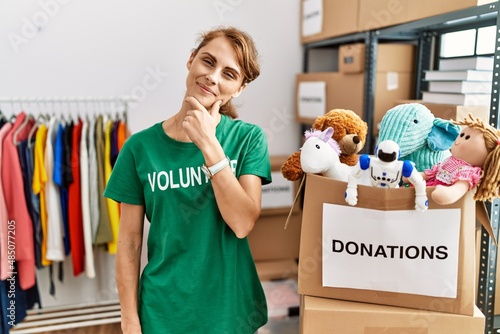 Beautiful caucasian woman wearing volunteer t shirt at donations stand looking confident at the camera smiling with crossed arms and hand raised on chin. thinking positive.