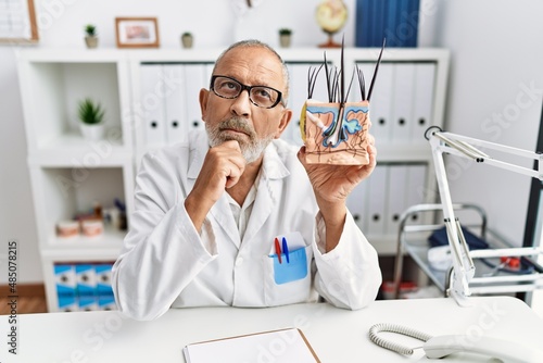 Mature doctor man holding model of human anatomical skin and hair serious face thinking about question with hand on chin  thoughtful about confusing idea