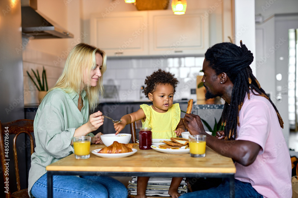 Beautiful family having breakfast together, enjoy meal at home in kitchen, in the morning. Attractive blonde mother, black dad and black daughter sit behind table eating food. everyday lifestyle
