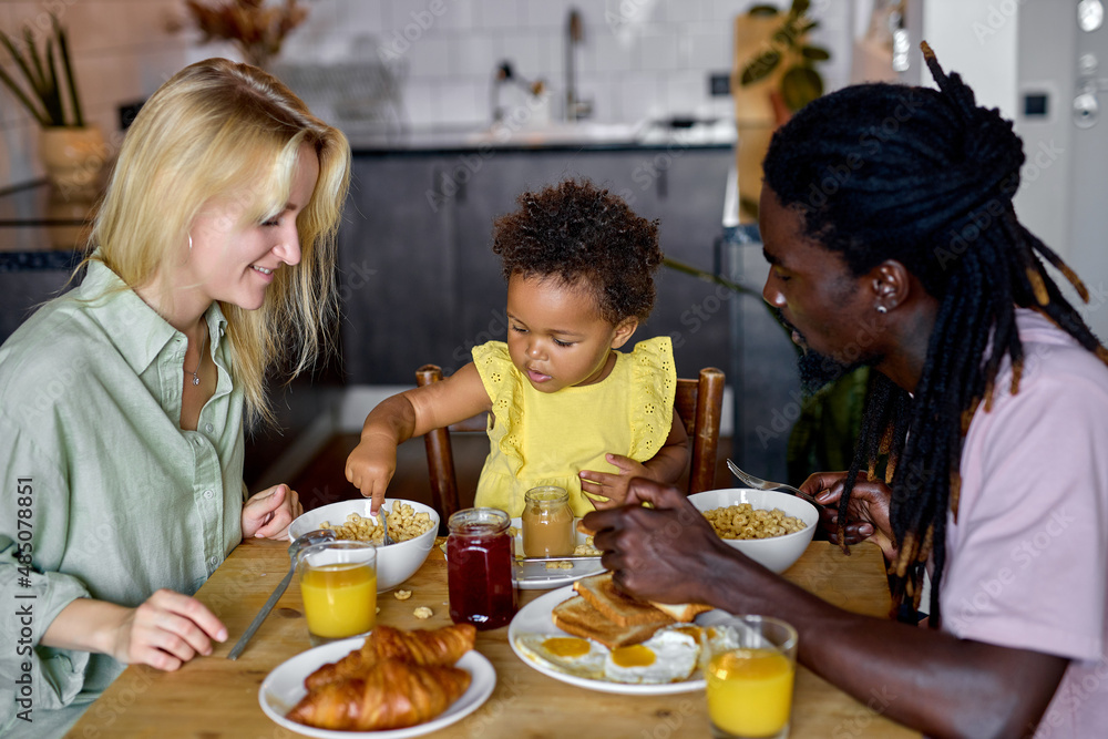 Little black child girl sitting in high chair and eating food with parents indoors. baby's first feeding. cute toddler holding spoon. in the morning, in cozy kitchen. diverse man and woman with child