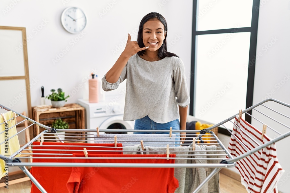 Obraz premium Young hispanic woman putting fresh laundry on clothesline smiling doing phone gesture with hand and fingers like talking on the telephone. communicating concepts.