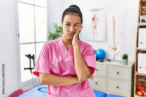 Young hispanic woman working at pain recovery clinic thinking looking tired and bored with depression problems with crossed arms.