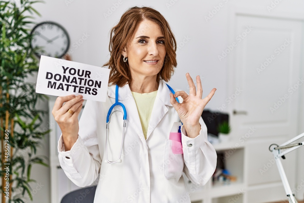 Middle age doctor woman holding your donation matters at the clinic doing ok sign with fingers, smiling friendly gesturing excellent symbol Middle age doctor woman holding your donation matters at the clinic doing ok sign with fingers, smiling friendly gesturing excellent symbol