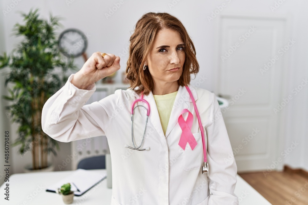 Middle age doctor woman wearing pink cancer ribbon on uniform strong person showing arm muscle, confident and proud of power Middle age doctor woman wearing pink cancer ribbon on uniform strong person showing arm muscle, confident and proud of power