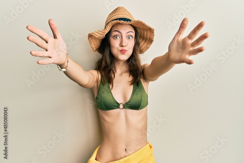 Young brunette woman wearing bikini and hat with open arms hugging puffing cheeks with funny face. mouth inflated with air, catching air.