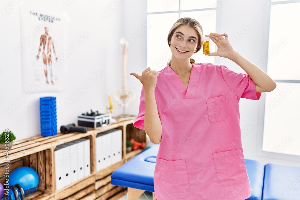 Young physiotherapist woman working at pain recovery clinic holding pills pointing thumb up to the side smiling happy with open mouth