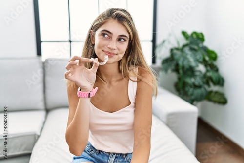 Young blonde woman holding invisible aligner orthodontic smiling looking to the side and staring away thinking.