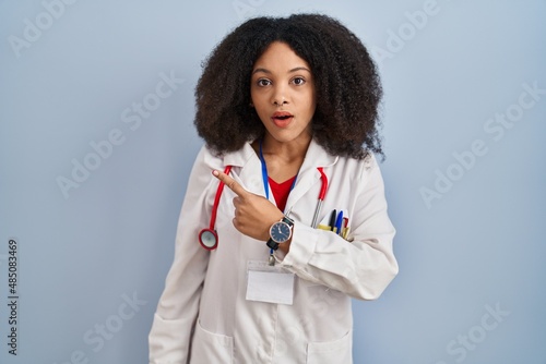 Young african american woman wearing doctor uniform and stethoscope surprised pointing with finger to the side  open mouth amazed expression.