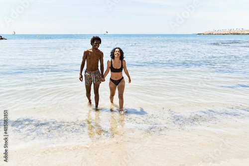 Young interracial tourist couple wearing swimwear walking with hands together at the beach.