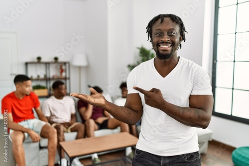Young african man with friends at the living room amazed and smiling to the camera while presenting with hand and pointing with finger.