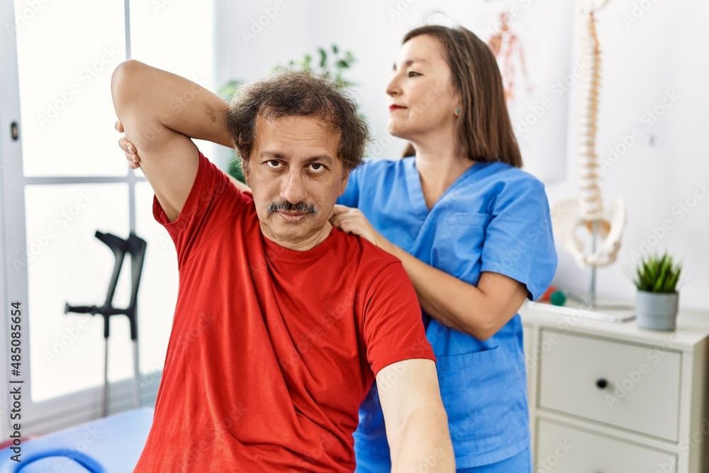 Obraz premium Middle age man and woman wearing physiotherapy uniform having rehab session stretching arm at physiotherapy clinic