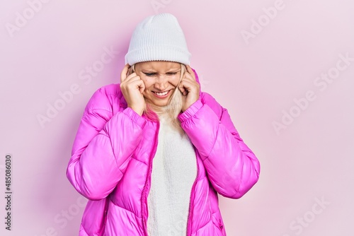 Beautiful caucasian blonde woman wearing wool hat and winter coat covering ears with fingers with annoyed expression for the noise of loud music. deaf concept. © Krakenimages.com