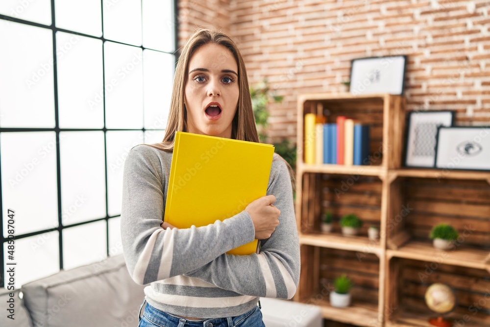 Young woman holding notebook in shock face, looking skeptical and sarcastic, surprised with open mouth Young woman holding notebook in shock face, looking skeptical and sarcastic, surprised with open mouth