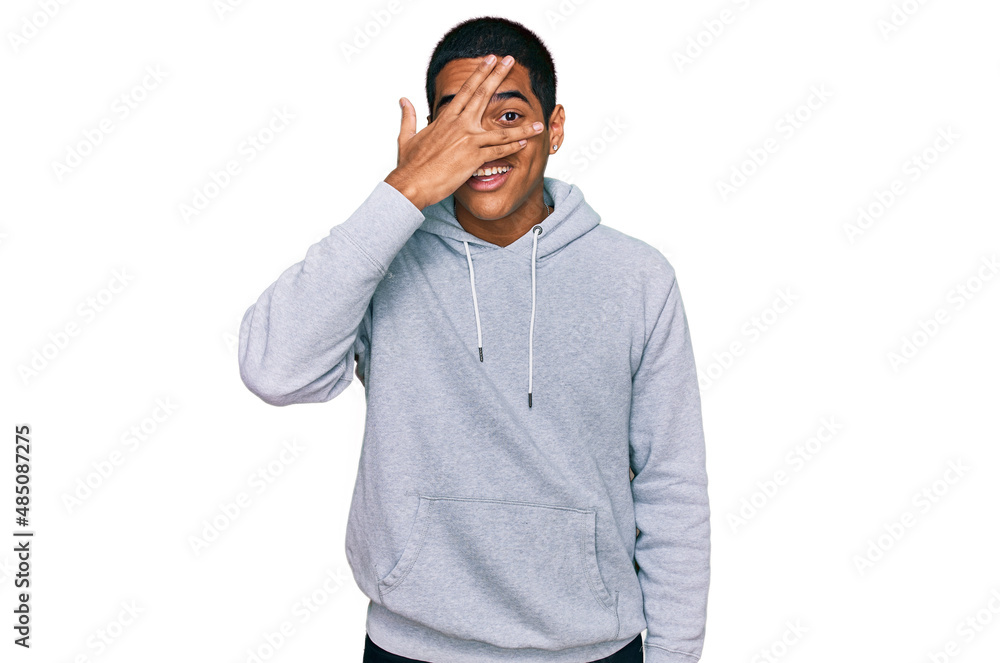 Young handsome hispanic man wearing casual sweatshirt peeking in shock covering face and eyes with hand, looking through fingers with embarrassed expression. Young handsome hispanic man wearing casual sweatshirt peeking in shock covering face and eyes with hand, looking through fingers with embarrassed expression.