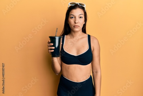 Young latin woman wearing bikini drinking soda thinking attitude and sober expression looking self confident