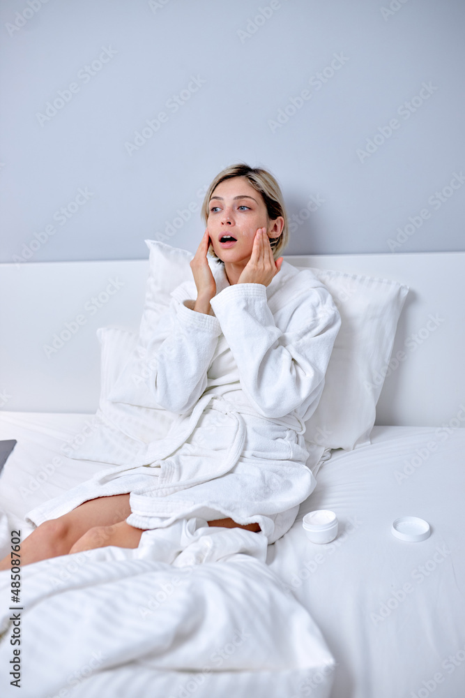 young woman in bathrobe applying cream on face touching moisturized soft healthy sensitive skin doing morning hydration spa beauty routine sitting on bed, facial skincare treatment concept young woman in bathrobe applying cream on face touching moisturized soft healthy sensitive skin doing morning hydration spa beauty routine sitting on bed, facial skincare treatment concept