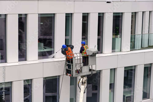 Two workers wearing safety harness wash office building facade at height standing in a crane cradle or aerial platform using pressure washer and mops