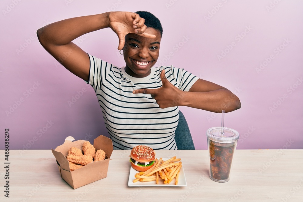 Young african american woman eating a tasty classic burger with fries and soda smiling making frame with hands and fingers with happy face. creativity and photography concept.