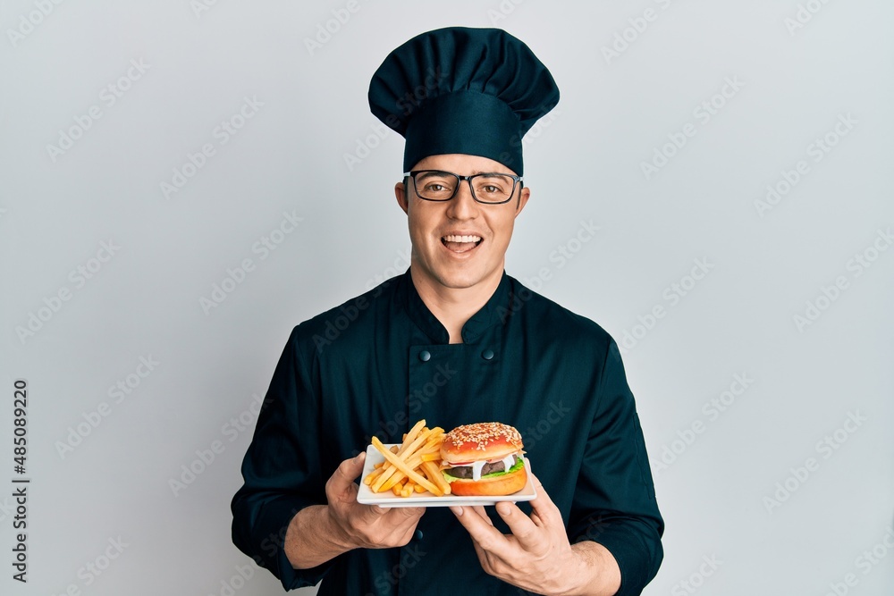 Handsome young man chef holding burger with fries celebrating crazy and amazed for success with open eyes screaming excited. Handsome young man chef holding burger with fries celebrating crazy and amazed for success with open eyes screaming excited.