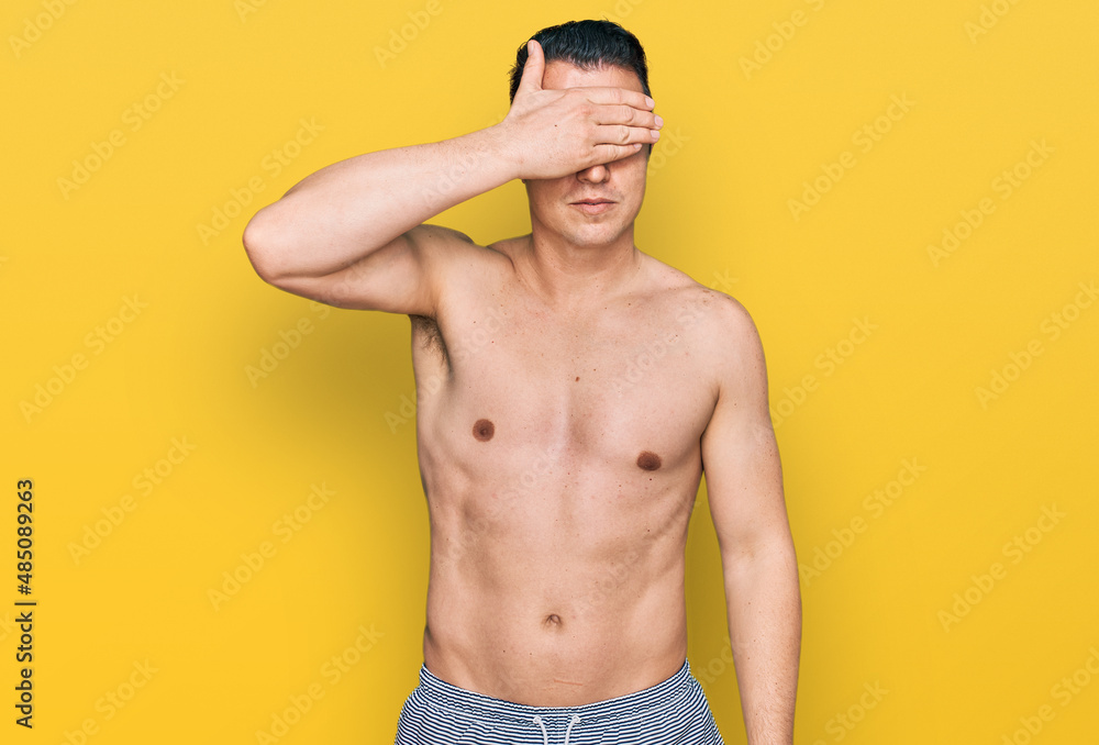 Handsome young man wearing swimwear shirtless covering eyes with hand, looking serious and sad. sightless, hiding and rejection concept