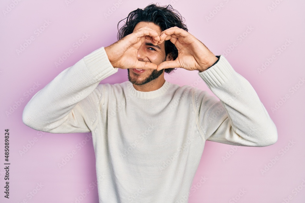 Handsome hispanic man wearing casual white sweater doing heart shape with hand and fingers smiling looking through sign Handsome hispanic man wearing casual white sweater doing heart shape with hand and fingers smiling looking through sign