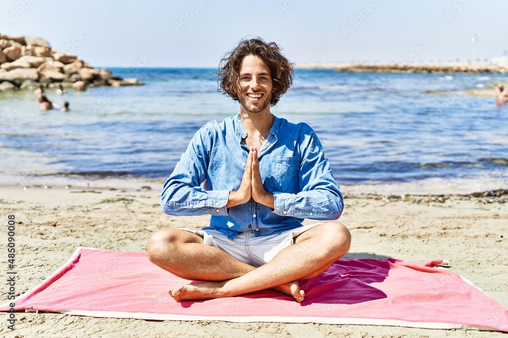 Young hispanic man relaxed doing yoga sitting on the sand at the beach.