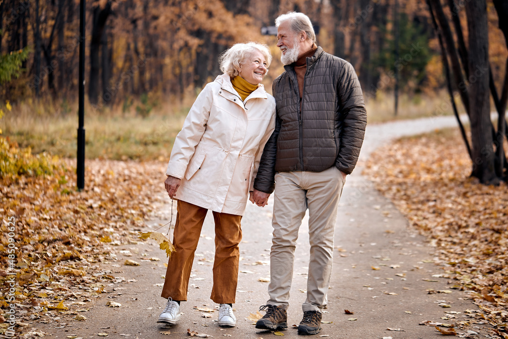 Fototapeta premium Happy elderly couple in love engaged in active walking in park, autumn season. Aged caucasian european gray haired man and woman in casual wear having fun, laughing together, dressed casually.