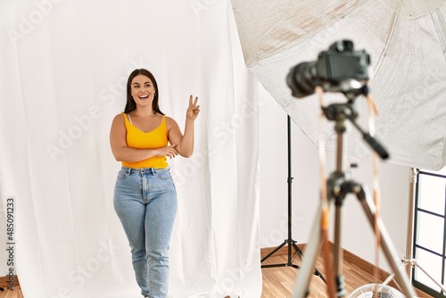 Young beautiful hispanic woman posing as model at photography studio smiling with happy face winking at the camera doing victory sign. number two.
