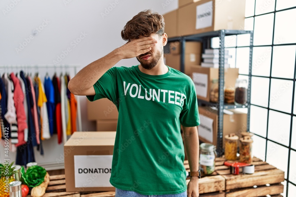 Young arab man wearing volunteer t shirt at donations stand covering eyes with hand, looking serious and sad. sightless, hiding and rejection concept