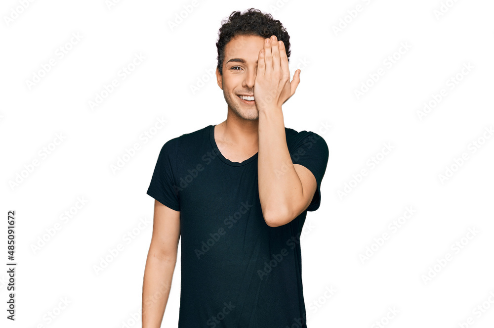 Young handsome man wearing casual black t shirt covering one eye with hand, confident smile on face and surprise emotion. Young handsome man wearing casual black t shirt covering one eye with hand, confident smile on face and surprise emotion.